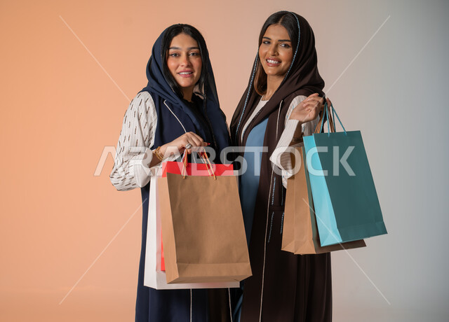 النظر إلى الكاميرا بإيماءات السعادة والسرور، شراء المستلزمات والهدايا، قضاء أوقات ممتعة في التسوق رفقة الأصدقاء، بورتريه مقرب لشابتين عربيتين خليجيتين سعوديتين محجبتين ترتديان العباءات الخليجية تضعان نظارات الشمس وتحملان العديد من الأكياس الورقية، خلفية ملونة