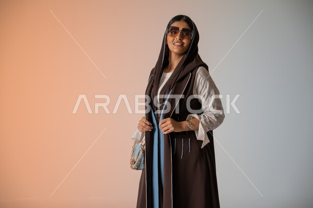 النظر إلى الكاميرا بإيماءات السعادة والسرور، بورتريه مقرب لشابة عربية خليجية سعودية محجبة ترتدي اللباس الكاجوال الساتر تقف وتمسك بالحقيبة الجلدية الفاخرة، مواكبة الموضة في الأزياء النسائية للمحجبات، الإكسسوارات والزينة والنظارات الشمسية المعاصرة، خلفية ملونة