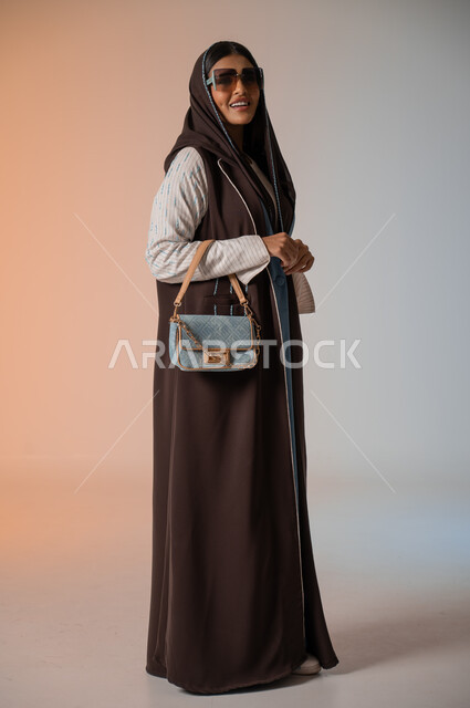 النظر إلى الكاميرا بإيماءات السعادة والسرور، بورتريه لشابة عربية خليجية سعودية محجبة ترتدي اللباس الكاجوال الساتر تقف وتمسك بالحقيبة الجلدية الفاخرة، مواكبة الموضة في الأزياء النسائية للمحجبات، الإكسسوارات والنظارات الشمسية المعاصرة، الجسم بالطول الكامل، خلفية ملونة