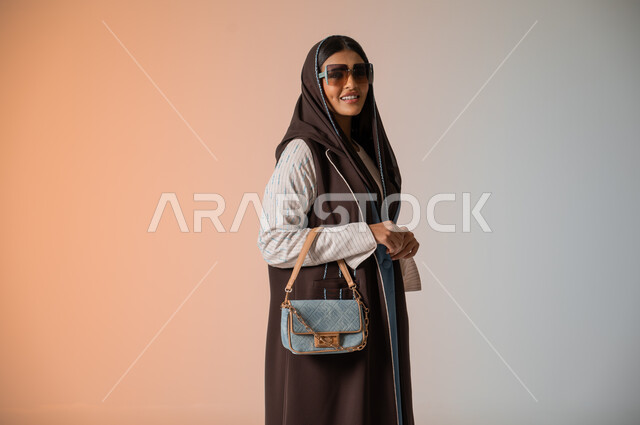 النظر إلى الكاميرا بإيماءات السعادة والسرور، بورتريه مقرب لشابة عربية خليجية سعودية محجبة ترتدي اللباس الكاجوال الساتر تقف وتمسك بالحقيبة الجلدية الفاخرة، مواكبة الموضة في الأزياء النسائية للمحجبات، الإكسسوارات والزينة والنظارات الشمسية المعاصرة، خلفية ملونة