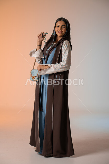 Looking at the camera with gestures of happiness and pleasure, portrait of a veiled Saudi Gulf Arab young woman wearing a casual, covering dress, standing and holding the luxurious leather bag, keeping up with fashion in women’s fashion for veiled women, contemporary accessories and sunglasses, full-length body, colorful background