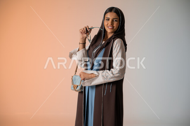 النظر إلى الكاميرا بإيماءات السعادة والسرور، بورتريه مقرب لشابة عربية خليجية سعودية محجبة ترتدي العباءة الخليجية العصرية تقف وتمسك بالحقيبة الجلدية الفاخرة، مواكبة الموضة في الأزياء النسائية للمحجبات، الإكسسوارات والزينة والنظارات الشمسية المعاصرة، خلفية ملونة