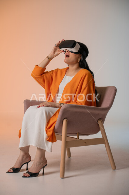 إيماءات التفاعل والاندماج مع الواقع الافتراضي، قضاء أوقات الفراغ بشكل ممتع، بورتريه لامرأة عربية خليجية سعودية أنيقة ترتدي الزي الكاجوال تجلس على كرسي مريح تضع نظارات الفي آر، استخدام الأجهزة التقنية الحديثة والتكنولوجيا في رؤية المشاهد الخيالية، خلفية ملونة