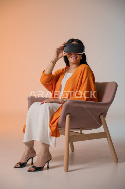 Gestures of interaction and integration with virtual reality, spending leisure time in a fun way, portrait of an elegant Saudi Arabian Gulf woman wearing casual clothes sitting on a comfortable chair wearing VR glasses, using modern technical devices and technology to see imaginary scenes, colorful background