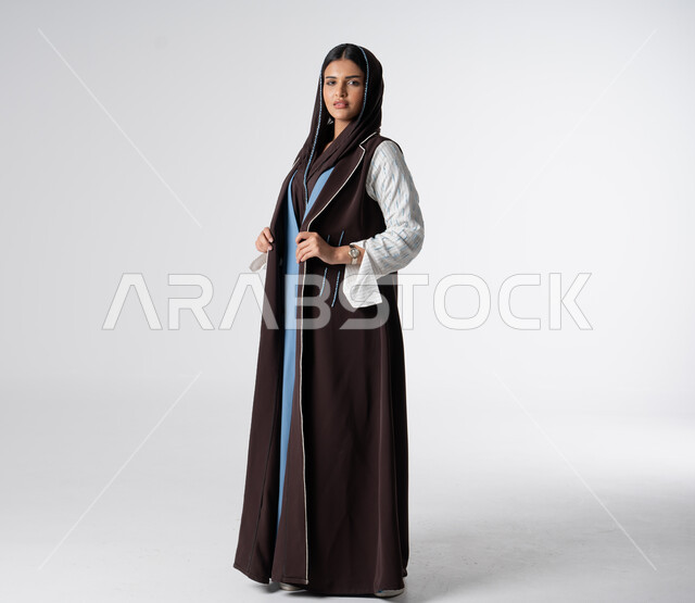 مفهوم الشياكة والاهتمام بالموضة, فن تصاميم العباءات العصرية الخليجية المميزة, أناقة النساء السعوديات, بورتريه من الجانب لعارضة أزياء عربية خليجية سعودية ترتدي العباءة والحجاب تنظر إلى الكاميرا بايماءات الثقة بالنفس، صورة الجسم بالطول الكامل، خلفية بيضاء