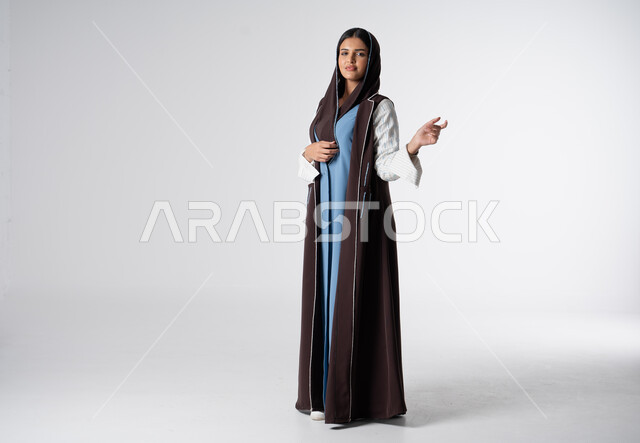 مفهوم الشياكة والاهتمام بالموضة, فن تصاميم العباءات العصرية الخليجية المميزة, أناقة النساء السعوديات, بورتريه من الجانب لعارضة أزياء عربية خليجية سعودية ترتدي العباءة والحجاب تنظر إلى الكاميرا بايماءات الثقة بالنفس، صورة الجسم بالطول الكامل، خلفية بيضاء