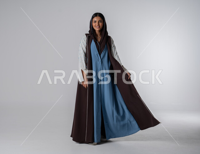 فن تصاميم العباءات العصرية الخليجية المميزة, موديلات أزياء للمحجبات, أناقة النساء السعوديات, بورتريه لإمرأة عربية خليجية سعودية ترتدي العباءة والحجاب تنظر إلى الكاميرا بايماءات الثقة بالنفس, مفهوم الشياكة والاهتمام بالموضة, صورة الجسم بالطول الكامل، خلفية