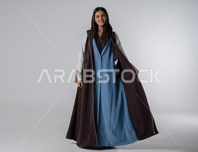 عباءات نسائية بتصاميم عصرية مميزة، بورتريه لإمرأه عربية سعوديه خليجية ترتدي عباءه ملونه أنيقه، مفهوم الموضة والاناقة، النظر إلى الكاميرا بايماءات الثقة بالنفس, الاهتمام بالمظهر الخارجي, صورة للجسم بالطول الكامل، خلفية بيضاء
