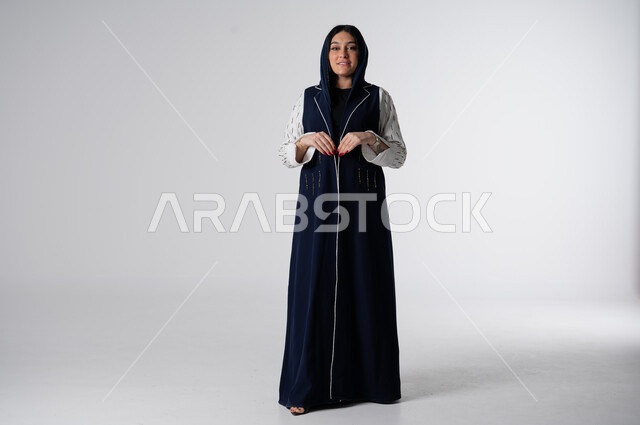 عباءات نسائية بتصاميم عصرية مميزة، بورتريه لإمرأه عربية سعوديه خليجية ترتدي عباءه ملونه أنيقه، مفهوم الموضة والاناقة، النظر إلى الكاميرا بايماءات الثقة بالنفس, الاهتمام بالمظهر الخارجي, صورة للجسم بالطول الكامل، خلفية بيضاء