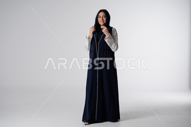 فن تصاميم العباءات العصرية الخليجية المميزة, موديلات أزياء للمحجبات, أناقة النساء السعوديات, بورتريه لإمرأة عربية خليجية سعودية ترتدي العباءة والحجاب تنظر إلى الكاميرا بايماءات الثقة بالنفس, مفهوم الشياكة والاهتمام بالموضة, صورة الجسم بالطول الكامل، خلفية بيضاء