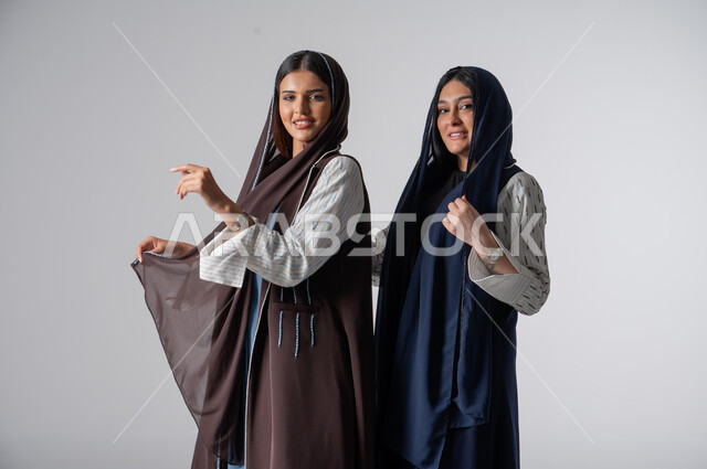 مفهوم العناية بالمظهر الخارجي، بورتريه لإمرأتين عربيتين خليجيتين سعوديتين ترتديان الحجاب والعباءة، قضاء أوقات ممتعة مع الأصدقاء، جلسة وناسة وتعارف، النظر الى الكاميرا بايماءات السعادة والسرور، خلفية بيضاء