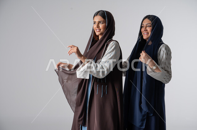 قضاء أوقات ممتعة مع الأصدقاء، مفهوم العناية بالمظهر الخارجي، بورتريه لإمرأتين عربيتين خليجيتين سعوديتين ترتديان الحجاب والعباءة ينظران إلى شيء ما، جلسة وناسة وتعارف، ايماءات السعادة والسرور، خلفية بيضاء