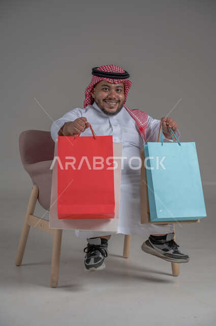 ذوي الإحتياجات الخاصة والهمم, بورتريه لشاب عربي خليجي سعودي يرتدي الثوب التقليدي والشماغ يحمل بيده مجموعة من الحقائب الملونة، الاعاقة الحركية وحمل اكياس التسوق، مفهوم الترفيه والتبضع, صورة الجسم بالطول الكامل، خلفية رمادية