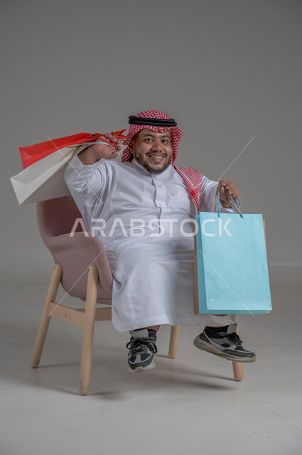 People with special needs and determination, a portrait of a Saudi Gulf Arab young man wearing a traditional thobe and shemagh, holding a group of colorful bags in his hand, mobility disability and carrying shopping bags, the concept of entertainment and shopping, full-length body portrait, gray background