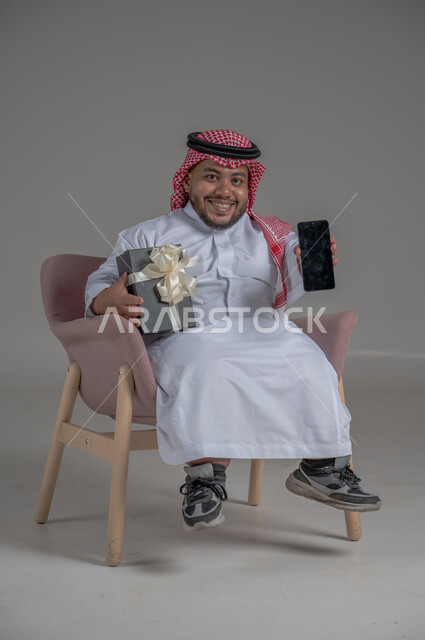 ذوي الإحتياجات الخاصة والهمم, مفهوم الهدايا والاعياد, بورتريه لشاب عربي خليجي سعودي يرتدي الثوب التقليدي والشماغ يجلس على كرسي متحرك ويحمل بيديه هدية, خلفية رمادية