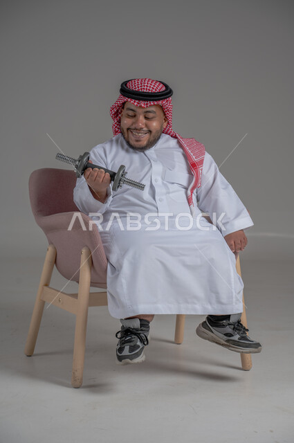 تنمية ودعم القدرة الحركية على حمل ورفع الأشياء، مفهوم الاهتمام بذوي الاحتياجات الخاصة، بورتريه لرجل عربي خليجي سعودي قزم يرتدي الثوب التقليدي والشماغ يجلس على كرسي  يحمل بيده دمبلز، صورة لكامل الجسم، خلفية رمادية