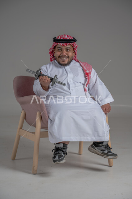 Developing and supporting the motor ability to carry and lift things, the concept of caring for people with special needs, a portrait of a dwarf Saudi Arabian Gulf man wearing a traditional thobe and shemagh, sitting on a chair holding dumbbells in his hand, full-body portrait, gray background