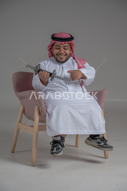 تنمية ودعم القدرة الحركية على حمل ورفع الأشياء، مفهوم الاهتمام بذوي الاحتياجات الخاصة، بورتريه لرجل عربي خليجي سعودي قزم يرتدي الثوب التقليدي والشماغ يجلس على كرسي  يحمل بيده دمبلز، صورة لكامل الجسم، خلفية رمادية