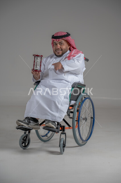 مفهوم إدارة الأعمال وتنظيم الأوقات، بورتريه لرجل سعودي خليجي قزم من ذوي الاحتياجات الخاصة يرتدي الثوب التقليدي والشماغ يجلس على كرسي متحرك يحمل بيده الساعة الرملية الزجاجية (البنكام)،  ايماءات السعادة والسرور, إدارة الجدول الزمني، خلفية رمادية