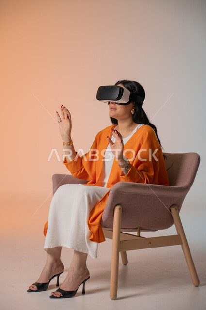قضاء أوقات الفراغ بشكل ممتع، إيماءات التفاعل والاندماج مع الواقع الافتراضي، بورتريه لامرأة عربية خليجية سعودية أنيقة ترتدي الزي الكاجوال تجلس على كرسي مريح تضع نظارات الفي آر، استخدام الأجهزة التقنية الحديثة والتكنولوجيا في رؤية المشاهد الخيالية، خلفية ملونة