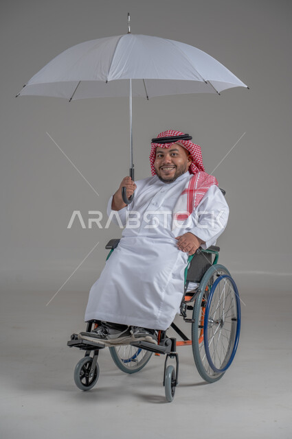 الحماية من الأمطار في الشتاء، دمج ذوي الهمم بالمجتمع السعودي، التظلل من أشعة الشمس، بورتريه لرجل عربي خليجي سعودي قزم مقعد يرتدي الثوب التقليدي والشماغ يجلس على كرسي متحرك يحمل بيده مظلة، صورة لكامل الجسم، خلفية رمادية