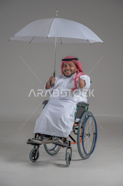 الحماية من الأمطار في الشتاء، دمج ذوي الهمم بالمجتمع السعودي، التظلل من أشعة الشمس، بورتريه لرجل عربي خليجي سعودي قزم مقعد يرتدي الثوب التقليدي والشماغ يجلس على كرسي متحرك يحمل بيده مظلة، صورة لكامل الجسم، خلفية رمادية