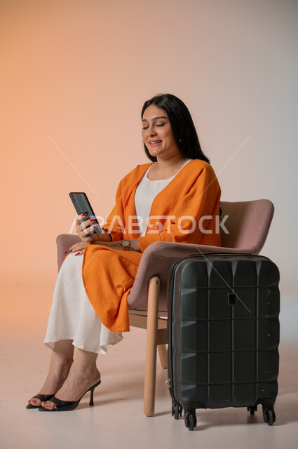 Integrating advanced technology into daily life, making video calls and communicating with family and friends, a portrait of an elegant Saudi Arabian Gulf woman wearing a casual outfit, sitting on a chair, holding a mobile phone in her hand and looking at it with gestures of pleasure, next to her is a travel bag, taking selfies, a colorful background.