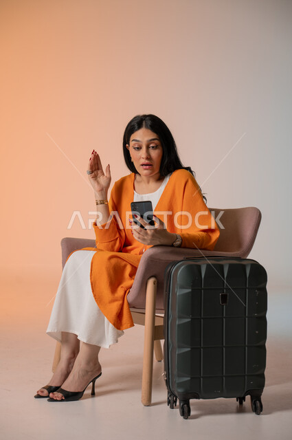 دمج التكنولوجيا المتطورة في الحياة اليومية، إجراء مكالمات الفيديو والتواصل مع الأهل والأصدقاء، بورتريه لامرأة أنيقة عربية خليجية سعودية ترتدي الزي الكاجوال تجلس على كرسي وتحمل بيدها الهاتف المحمول وتنظر إليه بإيماءات الدهشة  بجانبها شنطة السفر، خلفية ملونة