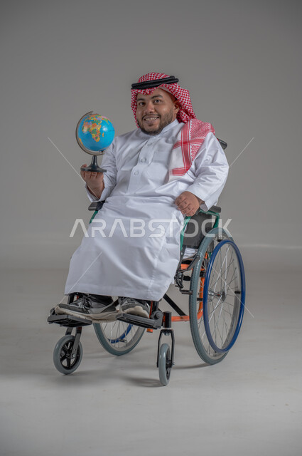 خريطة العالم الكروية, مفهوم البحث والاستكشاف حول العالم, بورتريه لرجل عربي خليجي سعودي  قزم مقعد من ذوي الهمم مبتسم يرتدي الثوب التقليدي والشماغ يحمل مجسم الكرة الارضية, ايماءات السعادة والسرور, صورة الجسم بالطول الكامل, خلفية رمادية
