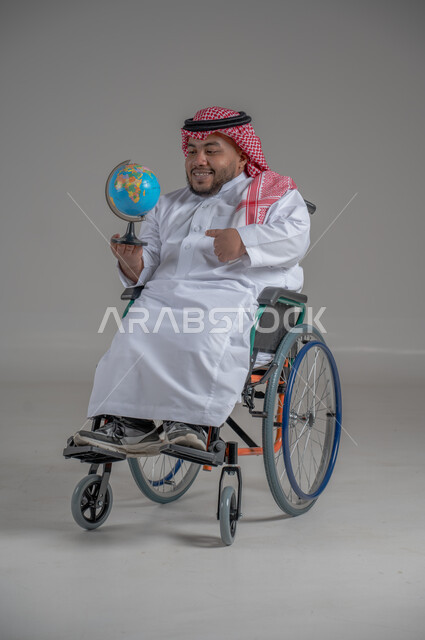 Spherical world map, concept of searching and exploring around the world, portrait of a smiling disabled dwarf Saudi Arabian Gulf man wearing traditional thobe and shemagh holding a globe, gestures of happiness and pleasure, full body image, gray background