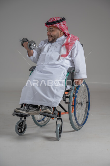 تنمية ودعم القدرة الحركية على حمل ورفع الأشياء، مفهوم الاهتمام بذوي الاحتياجات الخاصة، بورتريه لرجل عربي خليجي سعودي قزم مقعد يرتدي الثوب التقليدي والشماغ يجلس على كرسي متحرك يحمل بيده دمبلز، صورة لكامل الجسم، خلفية رمادية