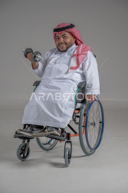 تنمية ودعم القدرة الحركية على حمل ورفع الأشياء، مفهوم الاهتمام بذوي الاحتياجات الخاصة، بورتريه لرجل عربي خليجي سعودي قزم مقعد يرتدي الثوب التقليدي والشماغ يجلس على كرسي متحرك يحمل بيده دمبلز، صورة لكامل الجسم، خلفية رمادية