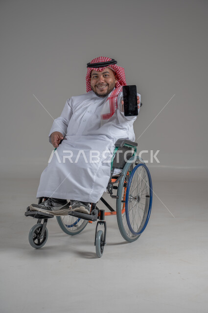 Mobile phone display with blank black screen, using modern advanced technical devices, looking at the camera with expressions of pleasure and happiness, portrait of a smiling dwarf Saudi Arabian Gulf man wearing a traditional keffiyeh and thobe holding a mobile phone in his hand, gray background