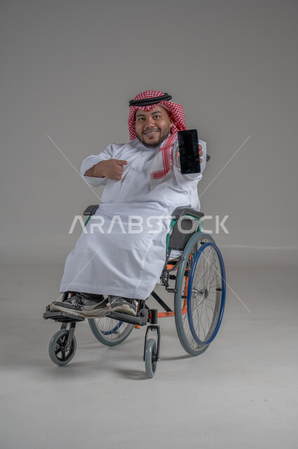 Mobile phone display with blank black screen, using modern advanced technical devices, looking at the camera with expressions of pleasure and happiness, portrait of a smiling dwarf Saudi Arabian Gulf man wearing a traditional keffiyeh and thobe holding a mobile phone in his hand, gray background