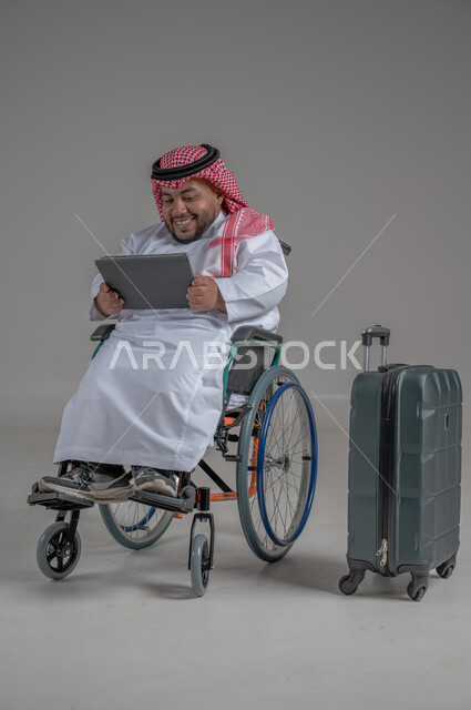 قضاء وقت الإجازة في السياحة والسفر، مفهوم الاسترخاء والاستراحة, بورتريه لولد عربي خليجي سعودي مقعد قزم يرتدي الثوب التقليدي والشماغ يجلس بجانب حقيبة السفر ويحمل بيده جهاز التابليت, ايماءات السعادة والسرور، صورة الجسم بالطول الكامل, خلفية رمادية