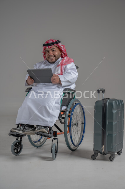 قضاء وقت الإجازة في السياحة والسفر، مفهوم الاسترخاء والاستراحة, بورتريه لولد عربي خليجي سعودي مقعد قزم يرتدي الثوب التقليدي والشماغ يجلس بجانب حقيبة السفر ويحمل بيده جهاز التابليت, ايماءات السعادة والسرور، صورة الجسم بالطول الكامل, خلفية رمادية