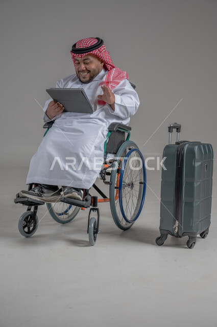 قضاء وقت الإجازة في السياحة والسفر، مفهوم الاسترخاء والاستراحة, بورتريه لولد عربي خليجي سعودي مقعد قزم يرتدي الثوب التقليدي والشماغ يجلس بجانب حقيبة السفر ويحمل بيده جهاز التابليت, ايماءات السعادة والسرور، صورة الجسم بالطول الكامل, خلفية رمادية