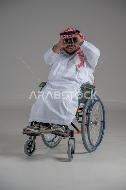 Using binoculars to detect distant objects, people with special needs and determination, portrait of a young Saudi Arabian Gulf dwarf in a wheelchair wearing traditional thobe and shemagh sitting in a wheelchair trying to see something, gray background