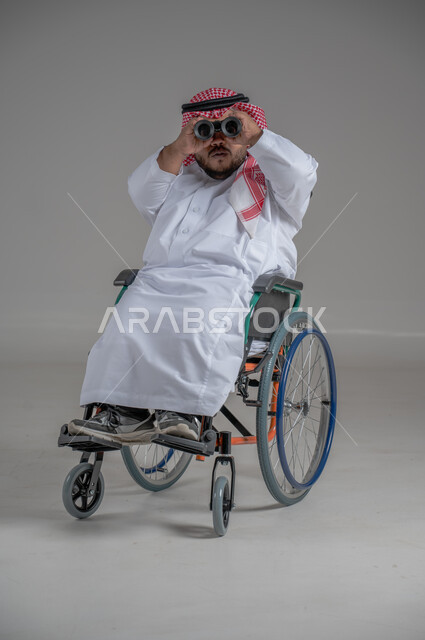 Using binoculars to detect distant objects, people with special needs and determination, portrait of a young Saudi Arabian Gulf dwarf in a wheelchair wearing traditional thobe and shemagh sitting in a wheelchair trying to see something, gray background