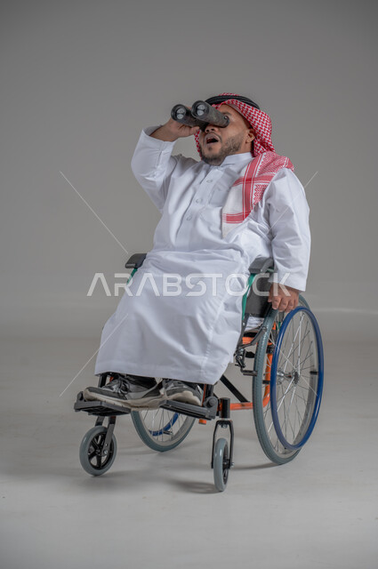 Using binoculars to detect distant objects, people with special needs and determination, portrait of a young Saudi Arabian Gulf dwarf in a wheelchair wearing traditional thobe and shemagh sitting in a wheelchair trying to see something, gray background
