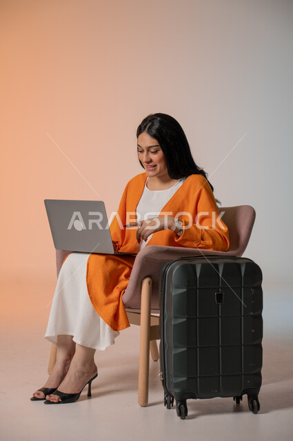 استخدام الاجهزة الحديثة والتقنية, دمج التكنولوجيا المتطورة في الحياة اليومية، بورتريه لامرأة أنيقة عربية خليجية سعودية ترتدي الزي الكاجوال تجلس على كرسي وتستخدم الحاسوب المحمول وتنظر إلىه بإيماءات السعادة والسرور بجانبها شنطة السفر، خلفية ملونة