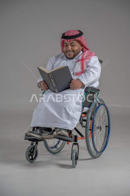 أسلوب حياة ذوي الهمم، دعم المجتمع السعودي لهوايات أصحاب الاحتياجات الخاصه، الاستمتاع بممارسة هواية المطالعة، بورتريه لرجل عربي خليجي سعودي قزم مقعد يرتدي الثوب التقليدي والشماغ يجلس على كرسي متحرك يقوم بقراءة كتاب، صورة لكامل الجسم، خلفية رمادية