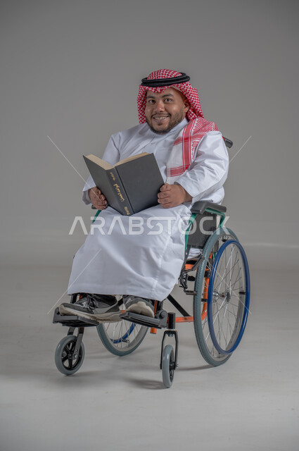 Lifestyle of people with disabilities, Saudi society support for hobbies of people with special needs, enjoying reading hobby, portrait of a Saudi Arabian Gulf dwarf man in wheelchair wearing traditional dress and shemagh sitting in a wheelchair reading a book, full body image, gray background