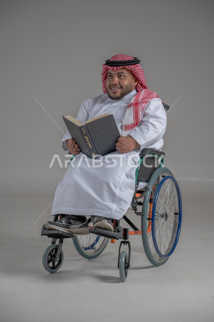 Lifestyle of people with disabilities, Saudi society support for hobbies of people with special needs, enjoying reading hobby, portrait of a Saudi Arabian Gulf dwarf man in wheelchair wearing traditional dress and shemagh sitting in a wheelchair reading a book, full body image, gray background