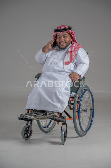 استخدام التكنولوجيا و التقنيات الحديثة، أسلوب حياة ذوي الهمم، الدردشة والتواصل مع الأهل والأصدقاء، بورتريه لرجل عربي خليجي سعودي قزم  مقعد يرتدي الثوب التقليدي والشماغ يجلس على كرسي متحرك يحمل بيده الهاتف المحمول، صورة لكامل الجسم، خلفية رمادية