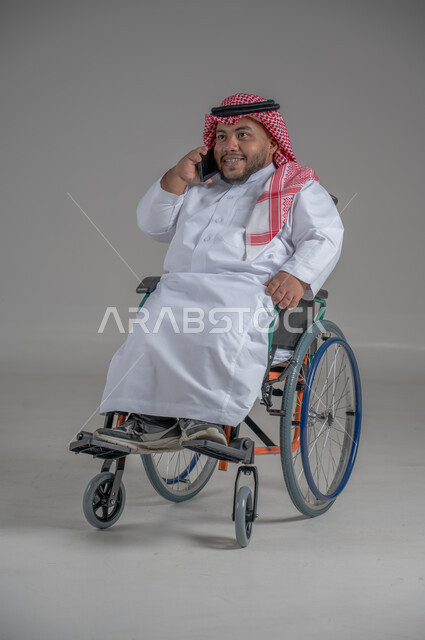استخدام التكنولوجيا و التقنيات الحديثة، أسلوب حياة ذوي الهمم، الدردشة والتواصل مع الأهل والأصدقاء، بورتريه لرجل عربي خليجي سعودي قزم  مقعد يرتدي الثوب التقليدي والشماغ يجلس على كرسي متحرك يحمل بيده الهاتف المحمول، صورة لكامل الجسم، خلفية رمادية