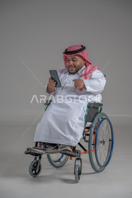 استخدام التكنولوجيا و التقنيات الحديثة، أسلوب حياة ذوي الهمم، الدردشة والتواصل مع الأهل والأصدقاء، بورتريه لرجل عربي خليجي سعودي قزم  مقعد يرتدي الثوب التقليدي والشماغ يجلس على كرسي متحرك يحمل بيده الهاتف المحمول، صورة لكامل الجسم، خلفية رمادية