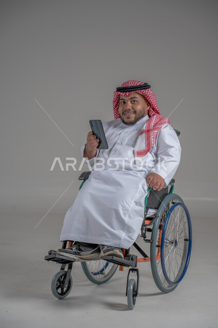 Use of technology and modern techniques, lifestyle of people with disabilities, chatting and communicating with family and friends, portrait of a disabled Saudi Arabian Gulf man wearing traditional thobe and shemagh sitting in a wheelchair holding a mobile phone in his hand, full body image, gray background