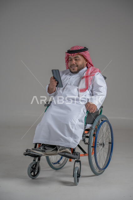 استخدام التكنولوجيا و التقنيات الحديثة، أسلوب حياة ذوي الهمم، الدردشة والتواصل مع الأهل والأصدقاء، بورتريه لرجل عربي خليجي سعودي قزم  مقعد يرتدي الثوب التقليدي والشماغ يجلس على كرسي متحرك يحمل بيده الهاتف المحمول، صورة لكامل الجسم، خلفية رمادية