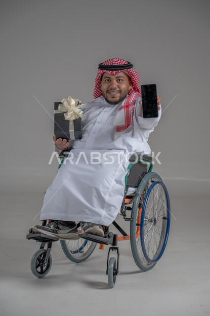 عرض شاشة سوداء فارغة، دعم المجتمع السعودي لذوي الهمم, مناسبات و أعياد سعيدة، بورتريه لرجل عربي خليجي سعودي قزم مقعد يرتدي الثوب التقليدي والشماغ يجلس على كرسي متحرك يحمل بيده صندوق هدايا والهاتف المحمول، شعور المعاقين بالحب و التقدير، صورة لكامل الجسم، خلفية رمادية